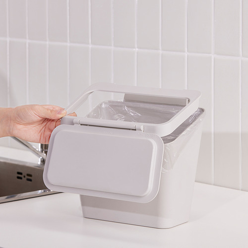 La persona abre la tapa del cubo de basura SKOLÄST, que cuelga en la puerta junto al fregadero. El cubo ayuda a mantener la cocina ordenada y evita los olores.