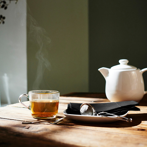 Taza de vidrio transparente con té humeante junto a una tetera blanca sobre una mesa de madera, con una cuchara y una servilleta cerca.