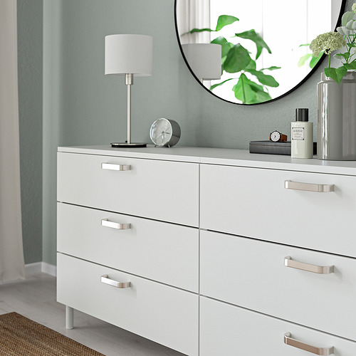 Cómoda blanca con seis cajones, tiradores modernos plateados. Rematado con una lámpara, un reloj y un perfume, reflejando una planta en un espejo redondo.