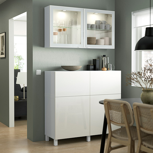 Moderno mueble BESTÅ contra pared verde con vitrinas, tazas. A la derecha, silla de mimbre y jarrón. Minimalista.