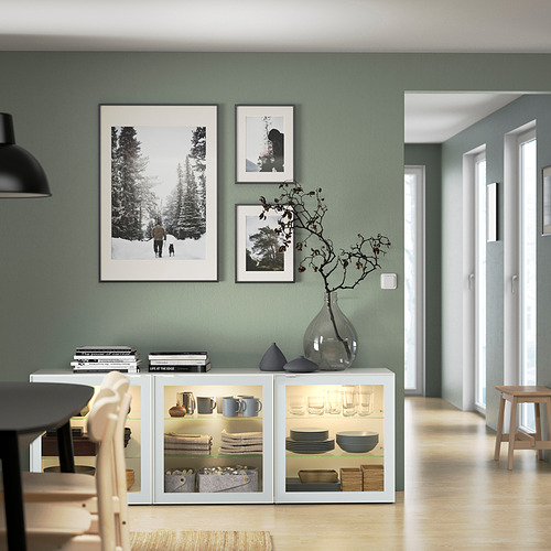 Comedor moderno con paredes de color verde salvia, aparador BESTÅ con vajilla y decoración, tres fotos enmarcadas.