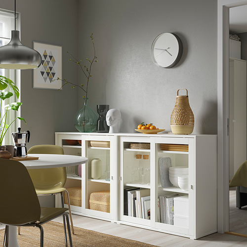 Comedor moderno con gabinete VIHALS blanco, puertas de vidrio y diseño elegante, que se ajusta entre las sillas y cerca de una pared gris.