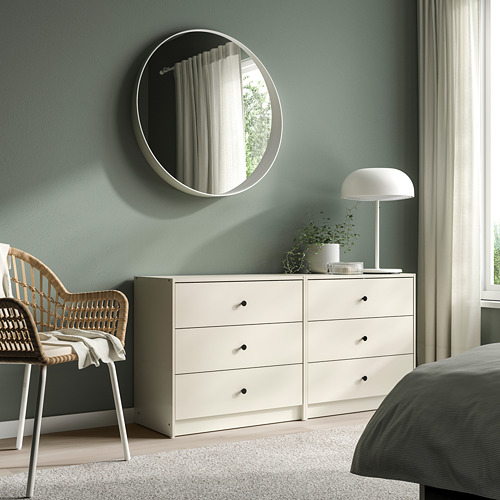 Dormitorio con cómoda blanca GURSKEN de 3 cajones al lado de la cama, cubierta con lámpara y planta, silla de mimbre, espejo redondo.