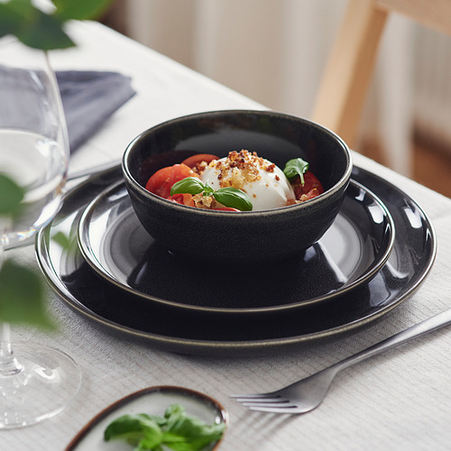 Tazón oscuro en platos apilados, sirviendo mozzarella, tomates y albahaca. Elegante mesa con vasos y cubiertos.
