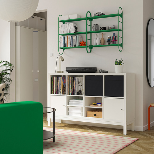 Estantería verde con libros, jarrones, cámaras sobre armario con vinilos, libros, impresora. Silla verde, alfombra a rayas debajo.