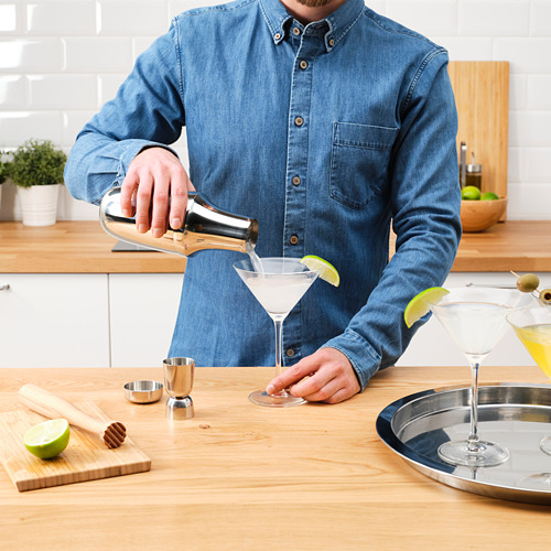 La persona vierte la bebida de la coctelera en una copa de martini con jigger y lima. El mostrador muestra herramientas e ingredientes para cócteles.