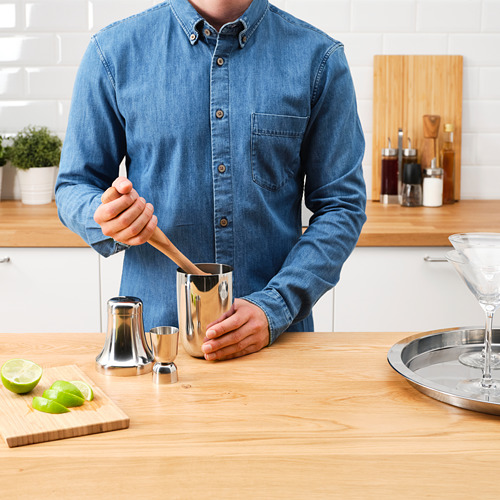 Un hombre con camisa de mezclilla usa una coctelera para preparar bebidas en un mostrador de madera. Incluye una coctelera, un jigger y una tabla de cortar con limas.