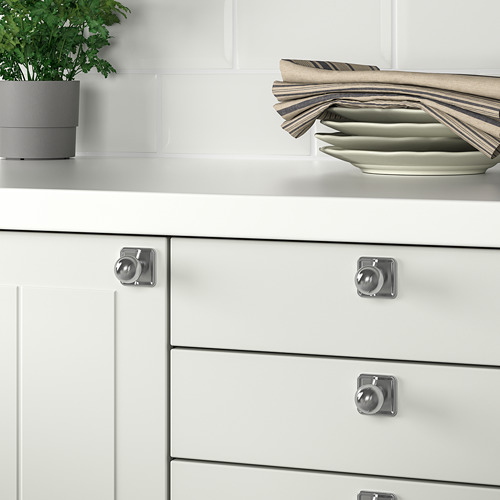 Cajones de cocina blancos con pomos y manijas plateadas, placas de acero inoxidable y una planta en maceta.