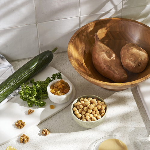 Cuenco de madera, platos pequeños con verduras, garbanzos y nueces sobre una encimera moteada de blanco, contra una pared de azulejos.