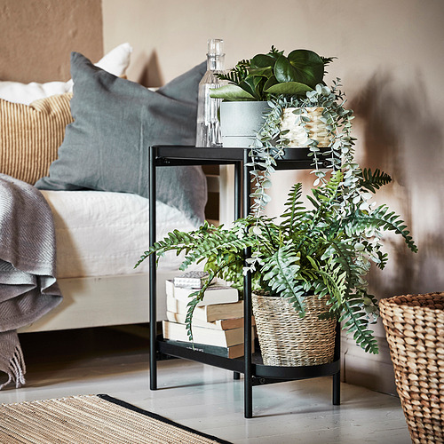 Moderno soporte de plantas de metal negro con cesta tejida, que sostiene plantas de hojas verdes, al lado de la cama con cojines y mantas.