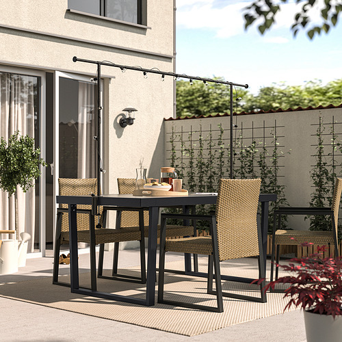 Patio moderno con mesa rectangular de metal negro, cuatro sillas de mimbre y guirnaldas de luces colgantes, ideal para cenar al aire libre o relajarse.