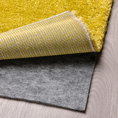 Tres alfombras superpuestas sobre un suelo de madera. La alfombra superior es amarilla con rayas azules y blancas, parte inferior de fieltro gris.