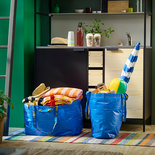 Dos grandes bolsas azules de IKEA FRAKTA llenas de artículos de playa, como toallas y juguetes, colocadas sobre una colorida alfombra frente a un armario verde.