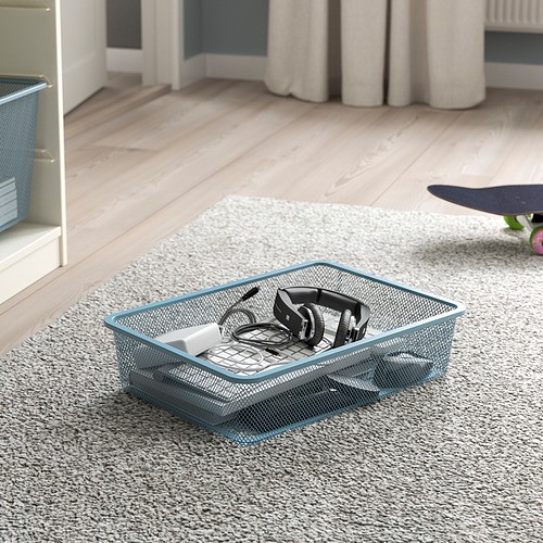 Cesta de almacenamiento de malla metálica sobre alfombra, con capacidad para auriculares y accesorios tecnológicos. Forma parte de la serie TROFAST de ikeas para organizar artículos.