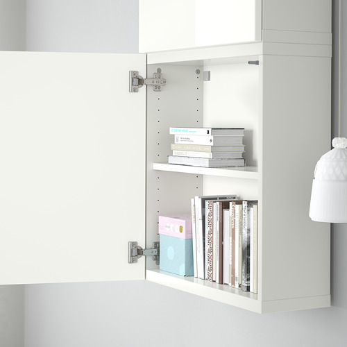 Armario blanco abierto con dos estantes, que contiene libros de colores y una caja azul. El armario se monta en la pared, formando parte del sistema de almacenamiento BESTÅ de ikea.