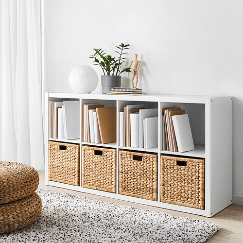 Estantería blanca kallax llena de libros y cestas tejidas. La parte superior sostiene la lámpara, la planta y la decoración. Las cestas almacenan los artículos de forma ordenada.