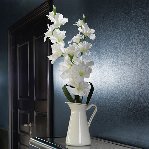 Jarrón esmaltado blanco SOCKERÄRT con flores blancas de tallo verde se encuentra en un estante oscuro contra una pared azul. Sencillo, elegante y luminoso.