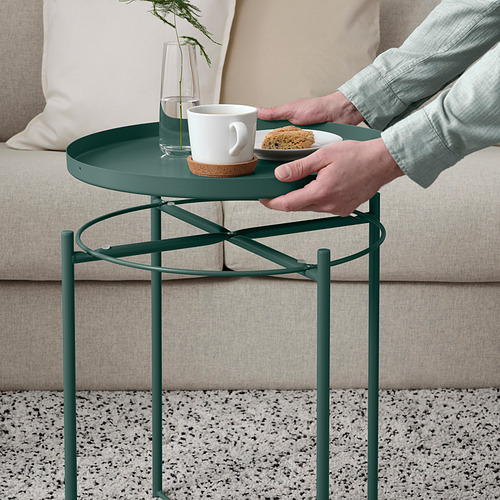 Persona sosteniendo una mesa de bandeja verde con taza de café y galleta, colocada al lado del sofá.
