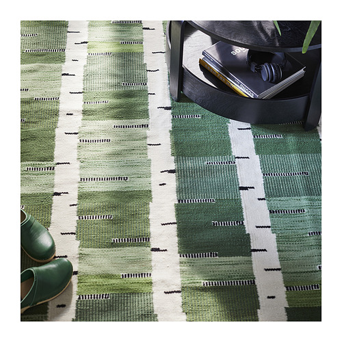 Alfombra de lana tejida a mano en verde y blanco con estampado de abedul. Reversible, de forma rectangular, colocada cerca de una mesa negra con artículos.
