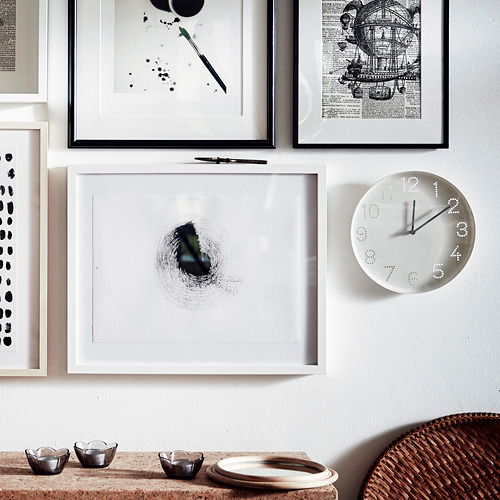 Marcos de arte de pared y un reloj blanco. La esfera del reloj muestra los números como puntos, las manecillas negras muestran la hora. Abajo, pequeños cuencos se colocan sobre una cesta tejida y una mesa.