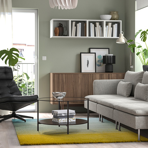 Sala de estar moderna con sofá gris, alfombra de colores, mesa de vidrio, gabinete de madera, silla de cuero, estantes con libros y paredes de color verde claro.