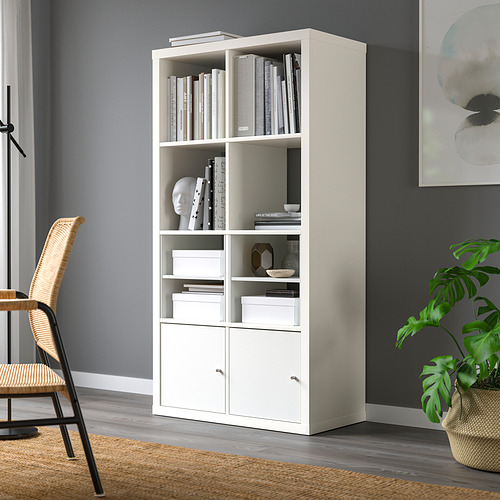 Estantería KALLAX blanca con libros, decoración y almacenamiento cerca de la pared gris. Silla de mimbre y planta cercana.
