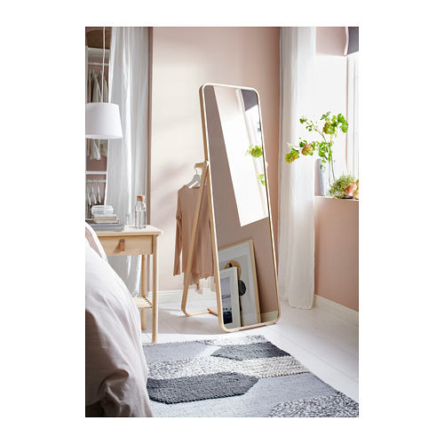 Dormitorio con cama blanca, mesa de madera y espejo de piso con marco dorado, perchero y ventana reflectantes.