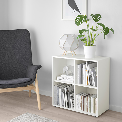 Salón moderno con librería EKET blanca, silla gris y elementos decorativos. Pequeña planta, lámpara única y libros ordenados en estanterías.