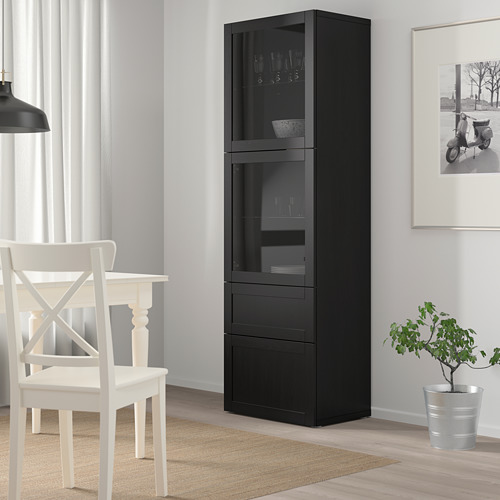 Mueble BESTÅ negro con puertas de cristal, con copas de vino y cuencos, junto a una silla blanca y una planta.