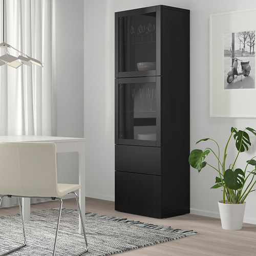 Un armario de almacenamiento BESTÅ negro con puertas de vidrio, más alto que una silla cercana, que exhibe copas de vino y platos, con una planta al lado.