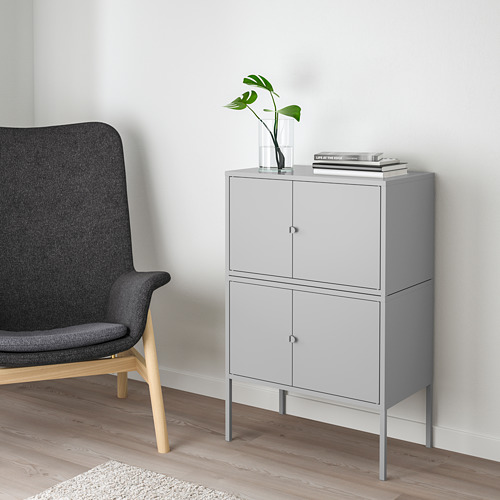 Mueble LIXHULT gris de 2 puertas, patas delgadas. Junto a una silla oscura, sostiene un jarrón de plantas y libros.