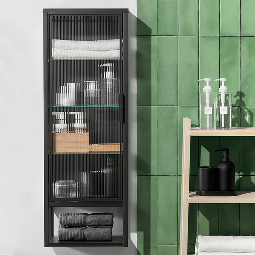 Mueble MOSSJÖN negro con estantes de cristal de caña, que contiene artículos de tocador, toallas. Diseño moderno con azulejos verdes.