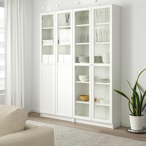 Gran estantería blanca BILLY con puertas de cristal, con platos y libros, de pie junto a una ventana con cortinas, junto a un sofá beige y una planta.
