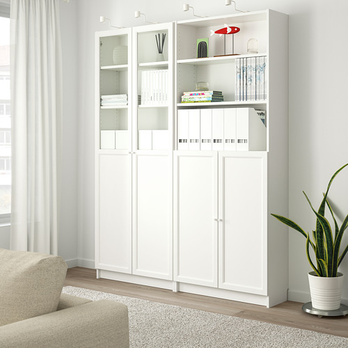 Librería blanca BILLY con puertas de cristal, llena de libros. Al lado de sillón beige y planta verde.
