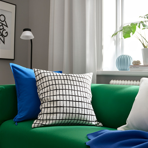 Sala de estar luminosa con sofá verde, almohadas azules, cojín a cuadros, lámpara blanca y plantas. La almohada MÄSTERROT en rejilla blanco/negro destaca por su lealtad.