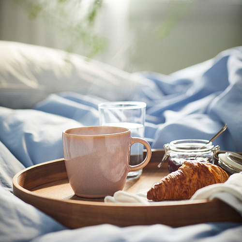 Bandeja de desayuno en la cama con taza, vaso, croissant y tarros de FÄRKKLAR. La taza es grande, beige, de cerámica con un diseño simple.