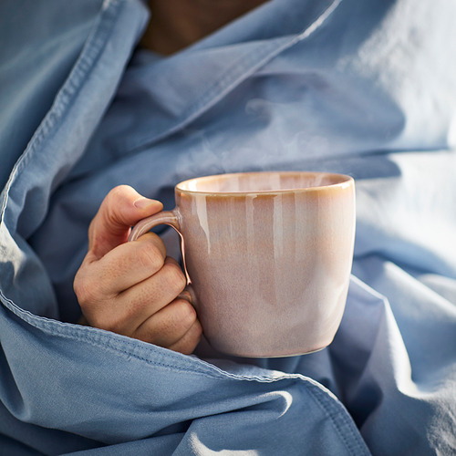 Mano sosteniendo una taza con vapor. La taza es de color claro, de diseño simple y diurno, adecuada para café o té. Envuelto en manta azul.