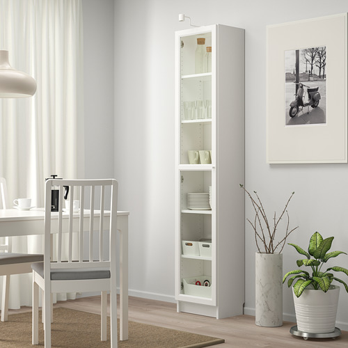 La estantería blanca con puerta de vidrio BILLY sostiene varios artículos de cocina, más alta que la silla de comedor circundante.