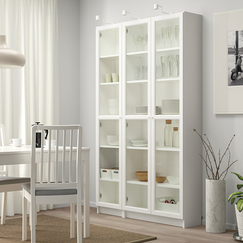 Librería blanca BILLY con puertas de cristal que exhiben platos, junto a mesa y sillas, jarrón en el suelo.