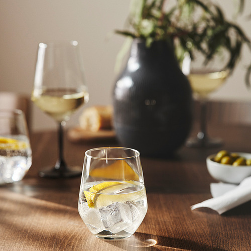 Sobre una mesa de madera se encuentra una copa de vino transparente con una rodaja de limón, junto a un jarrón de flores y un cuenco de aceitunas.