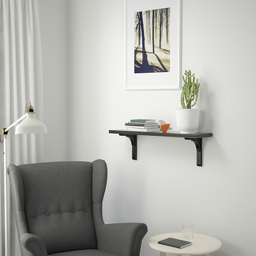 Una habitación acogedora con una silla gris y una estantería de pared BERGSHULT. La estantería contiene libros, una planta y una lámpara. El estante es más pequeño que la silla.