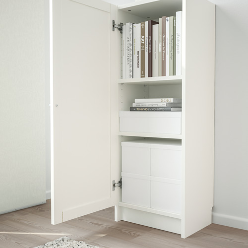 Librería blanca BILLY con una puerta abierta, exhibiendo estantes con libros y revistas, colocada sobre suelo de madera con puerta blanca en el fondo.