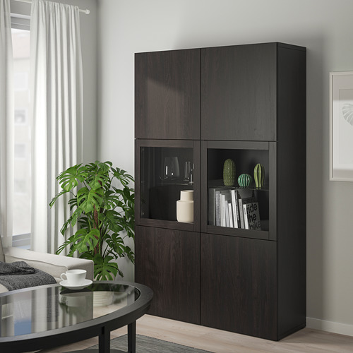Mueble de almacenamiento BESTÅ negro con puerta de cristal que exhibe jarrones y libros. Su gran tamaño encaja perfectamente en la sala de estar, junto a una mesa junto a una ventana.