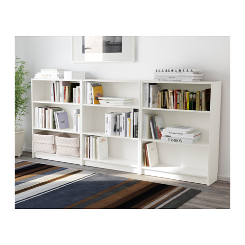 Estanterías blancas BILLY con estantes ajustables llenos de libros. Una planta y una alfombra a rayas debajo, una foto enmarcada cuelga encima.