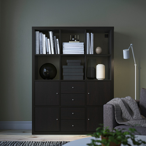 Estantería KALLAX negra contra pared verde, con varios objetos, junto a un sillón gris y una lámpara.