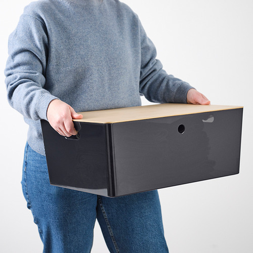 Persona que sostiene una caja rectangular gris claro con agujero, vestida con suéter azul y jeans.