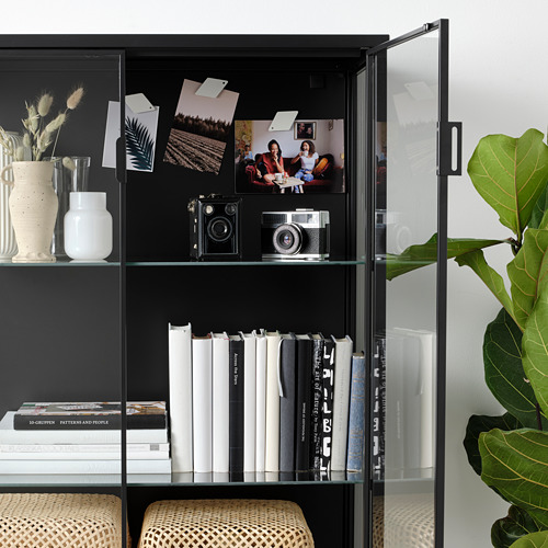 Mueble con puerta de cristal negro con estantes que muestran jarrones, cámaras, libros e imanes con fotos. Iluminación integrada y pies ajustables visibles.