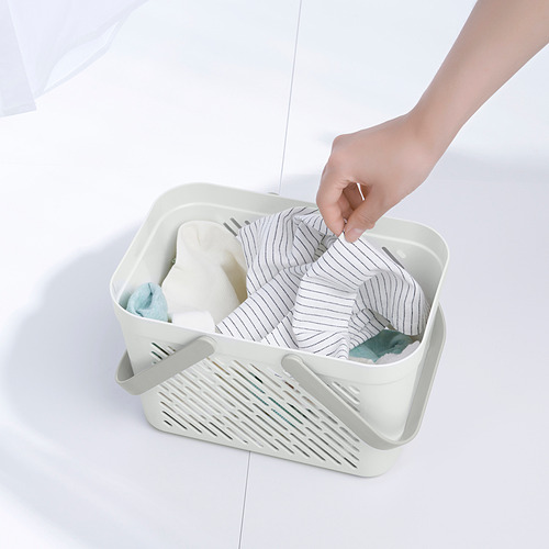La mano coloca el artículo en una cesta de almacenamiento RÅNEN blanca llena de toallas y artículos de tocador, mostrando su uso práctico para organizar los elementos esenciales del baño.