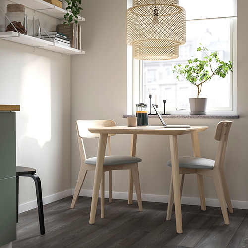 Mesa de comedor de madera LISABO con chapa de fresno, patas de abedul macizo y sillas a juego cerca de una ventana, además de un pequeño estante lateral y una lámpara colgante.