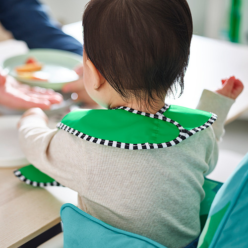 Un niño que lleva un babero verde impermeable con ribetes a cuadros se sienta en una silla alta, comiendo. El peto es ajustable y fácil de limpiar.